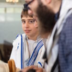 הנער מביט בעדינות לעבר המצלמה בעת קריאת התורה, בעוד הגברים לצידו מתרכזים בתפילין ובספר. תיעוד אותנטי של רגע אישי ומיוחד.