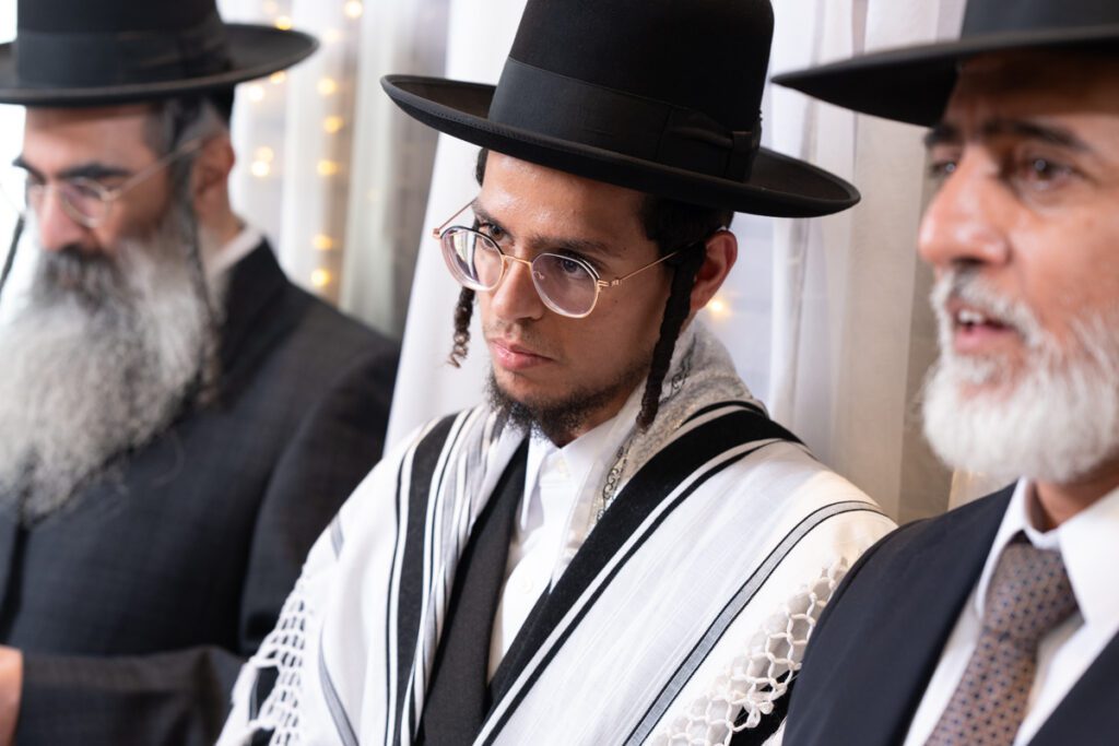 צילום חתונות חרדיות, גברים חרדים בלבוש חגיגי וטלית ברגע מרגש של תפילה ושירה בחתונה.