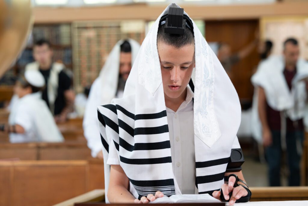 נער בר מצווה לבוש טלית ותפילין קורא בסידור בבית כנסת בתל אביב