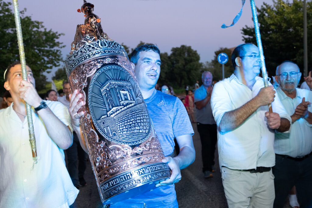 גבר עם חולצה כחולה נושא ספר תורה מהודר בתהלוכה בשעות הערב